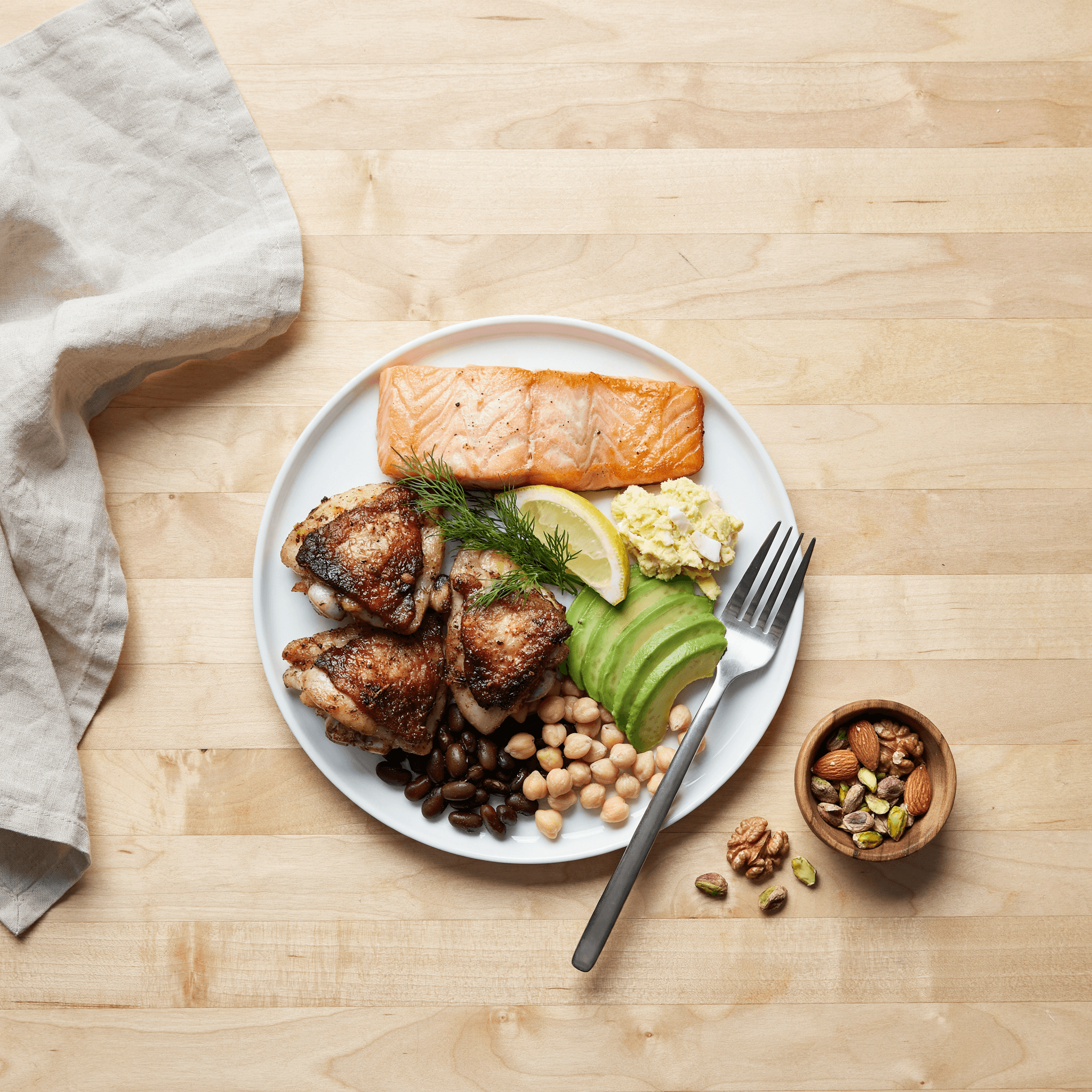 A healthy meal with salmon, roasted chicken, avocado, chickpeas, and nuts on a wooden table.