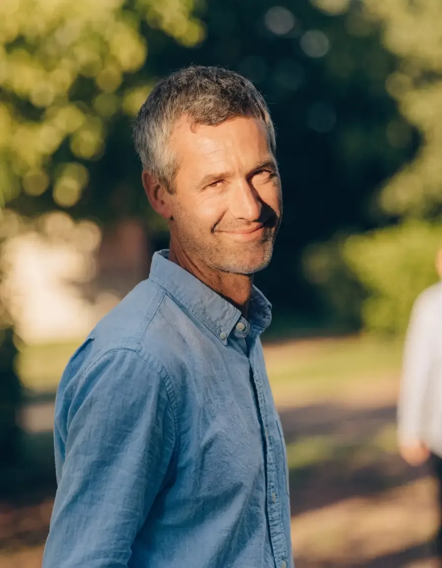 A man with graying hair and a blue shirt smiles subtly in an outdoor setting.